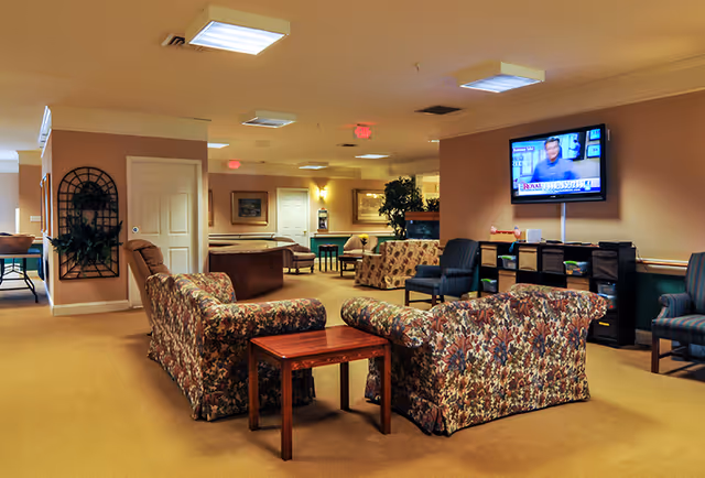 A spacious living room area in a senior living facility with floral patterned sofas, wooden side tables, and several armchairs. A flat-screen TV is mounted on the wall showing a news program. The room has beige walls, carpeted floors, and ceiling lights. There are plants and framed pictures decorating the space.