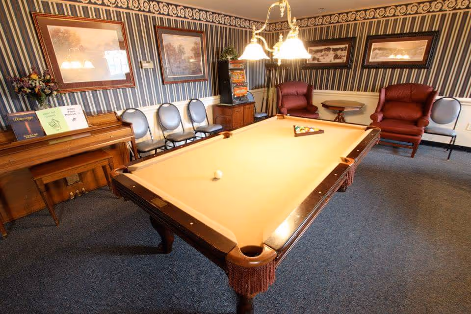 A recreational room with a pool table in the center, a piano with sheet music on top, several chairs lined up against the wall, two red armchairs, a small round table, and framed artwork on striped wallpapered walls.