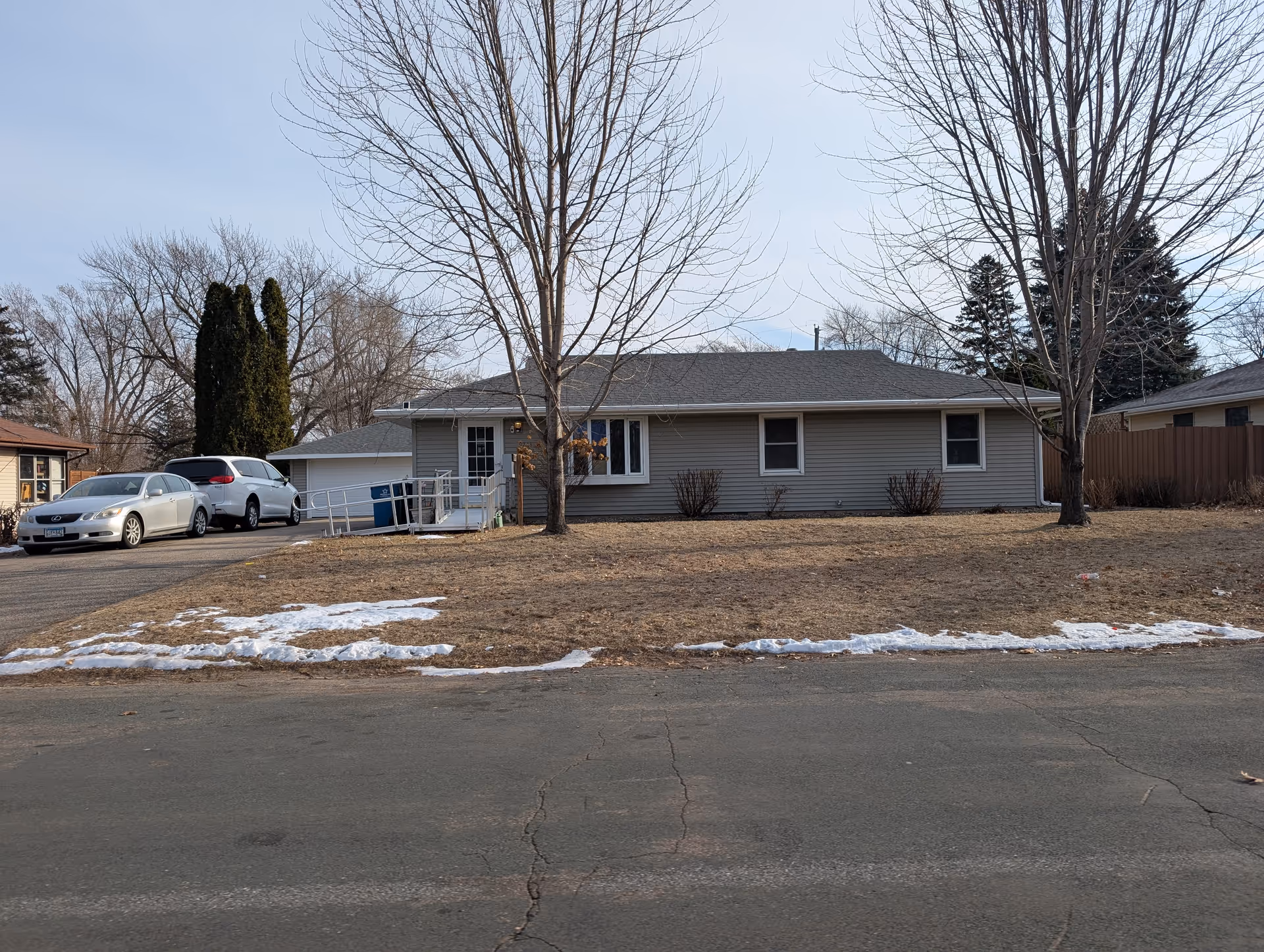 Single-story residential building with gray siding and a dark shingled roof, a driveway with two parked cars, a wheelchair ramp leading to the front door, leafless trees in the yard, and patches of snow on the ground.