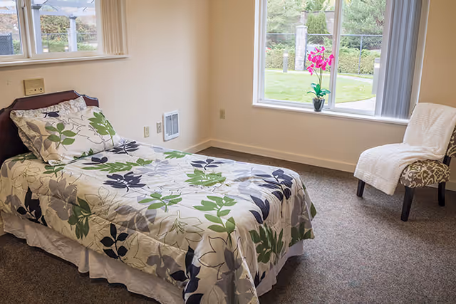 A simple bedroom with a single bed covered in a floral patterned bedspread in shades of green, gray, and black. There is a window with a view of greenery outside and a small potted plant with pink flowers on the windowsill. A cushioned chair with a white throw blanket is placed near the window.