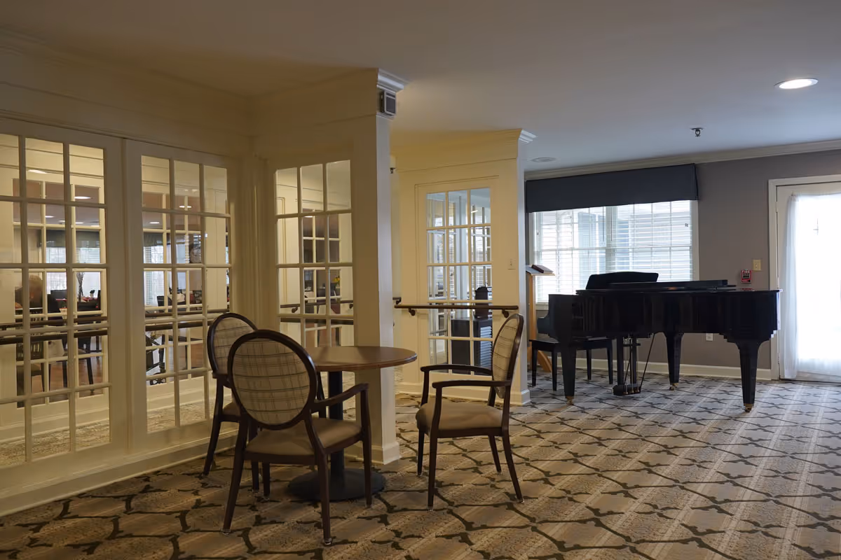 A cozy interior room with a round wooden table and four cushioned chairs with plaid backs. The room features large windows with white frames and a patterned carpet. A black grand piano is positioned near a window with blinds and a door with sheer curtains.