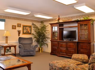 A cozy living room area in a senior living facility featuring a large wooden entertainment center with a television, a comfortable upholstered armchair, a patterned sofa, a wooden coffee table, a side table with a lamp, framed artwork on the walls, and a tall potted plant in the corner.