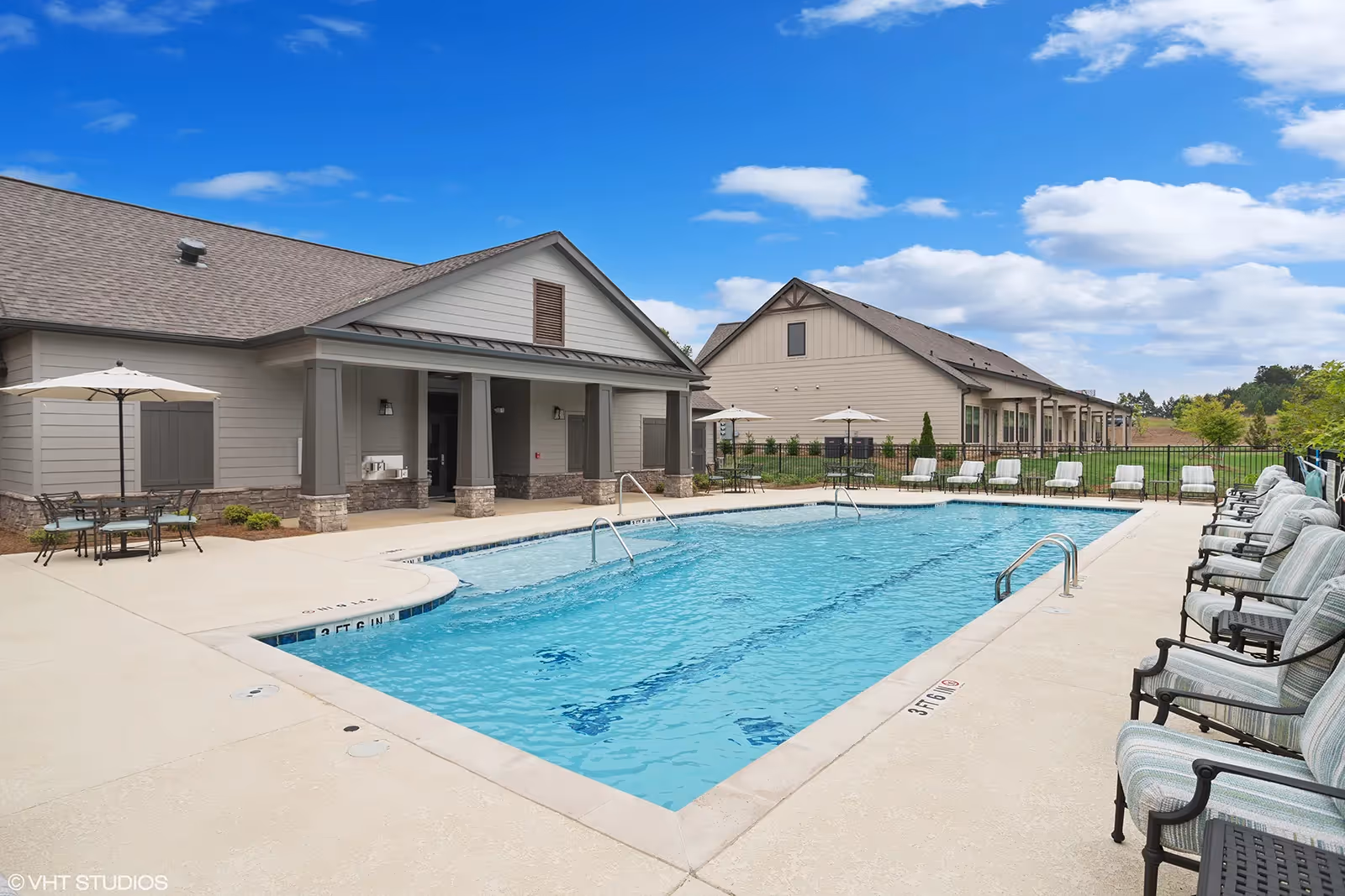 Outdoor swimming pool area with clear blue water surrounded by cushioned chairs and tables with umbrellas. Two beige buildings with gray roofs are visible in the background under a partly cloudy blue sky.