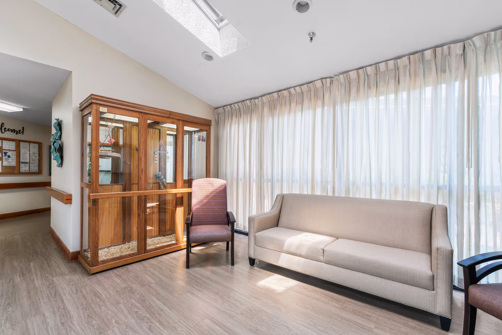 A bright sitting area in a nursing facility with a beige sofa and a purple armchair placed near large windows covered with sheer curtains. There is a wooden display cabinet with glass doors and a skylight above, allowing natural light to fill the space. The floor is covered with light wood laminate, and a hallway with a bulletin board is visible in the background.