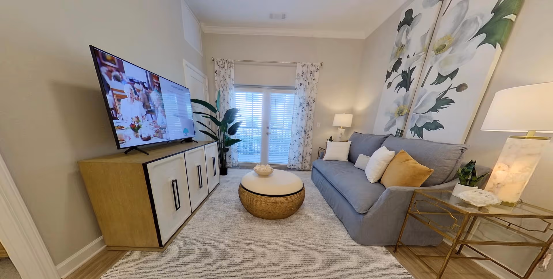 A cozy living room with a gray sofa adorned with white and mustard yellow pillows, a round woven ottoman in the center, a large flat-screen TV on a wooden cabinet, floral curtains on a glass door, and large floral artwork on the wall behind the sofa. There are two lamps on either side of the sofa and a potted plant near the door.