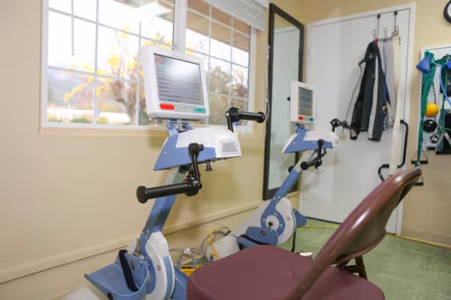 A small exercise room with two stationary exercise bikes facing a window. A chair with a maroon cushion is placed in front of the bikes. A large mirror is mounted on the wall behind the bikes, reflecting the equipment. Coats and exercise bands hang on hooks on a white door in the background.