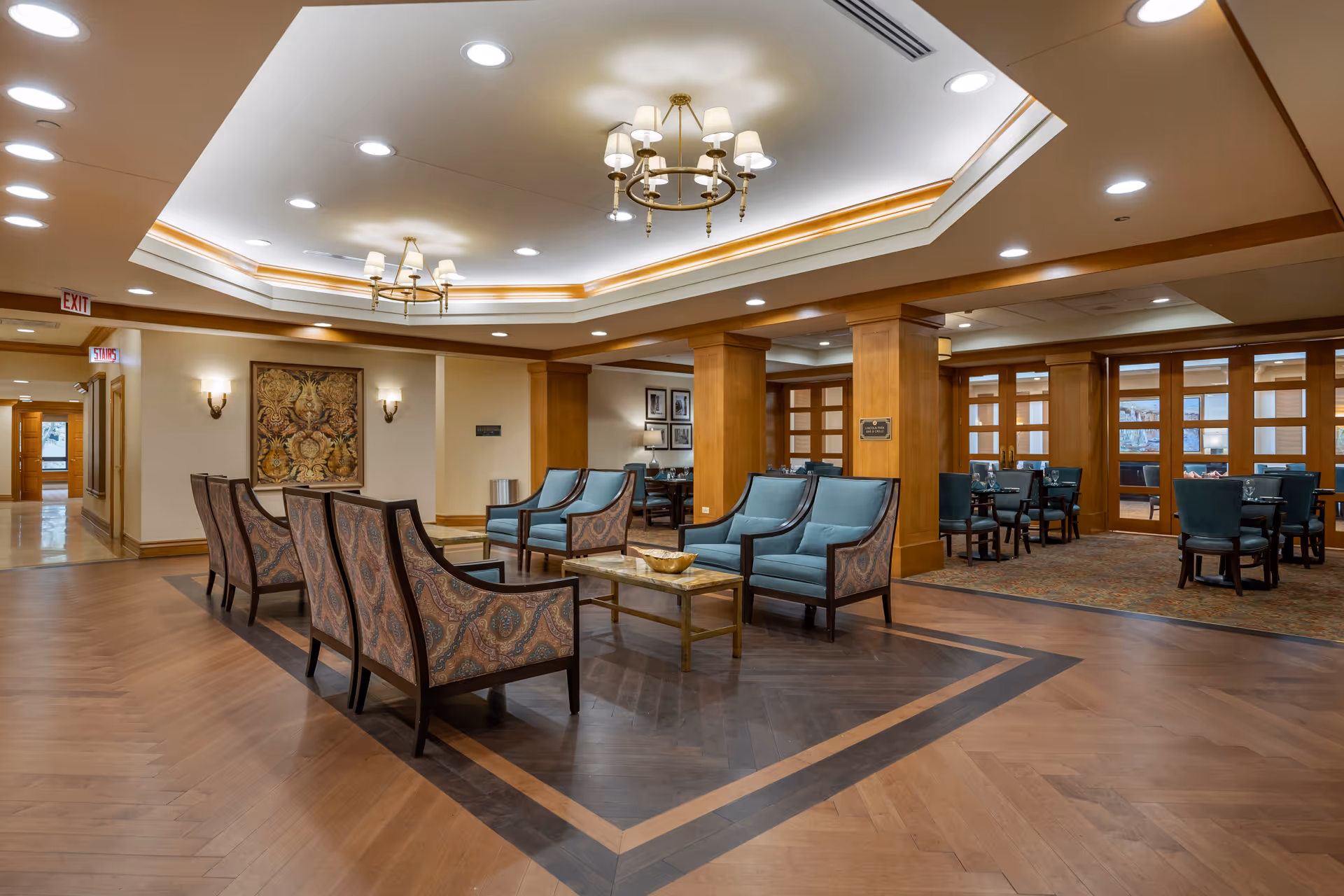 A spacious and well-lit common area in a senior living facility featuring a seating arrangement with patterned armchairs and blue cushioned chairs around marble-top coffee tables. The room has wooden flooring with a decorative inlay, wooden columns, and a ceiling with recessed lighting and chandeliers. In the background, there is a dining area with tables and chairs, separated by wooden-framed glass doors.