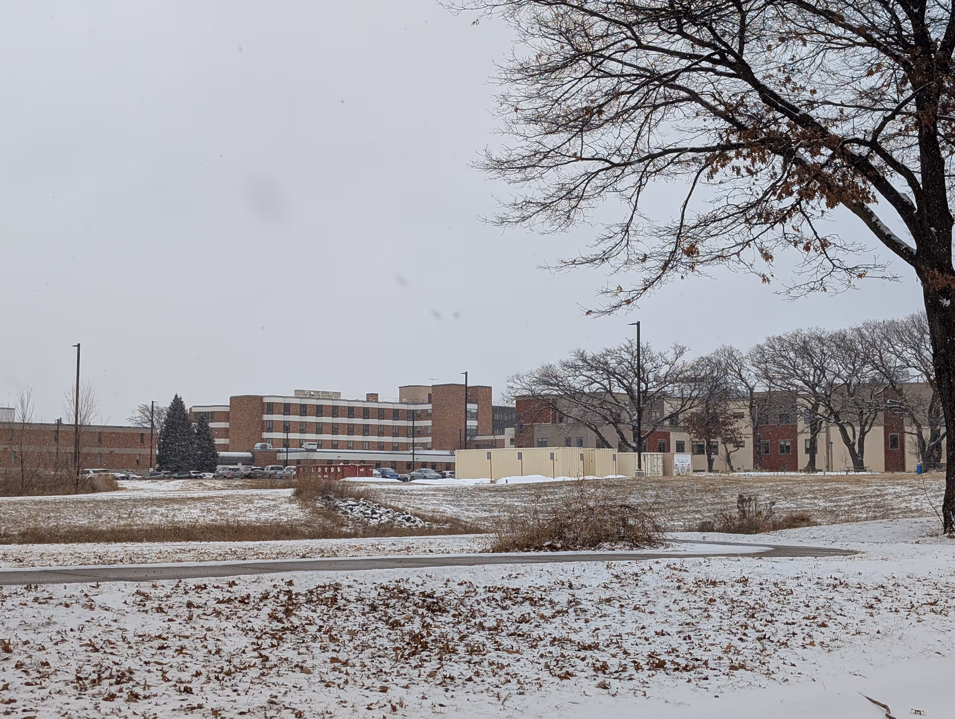 Snow-covered lawn and leaf-strewn foreground with a multi-story brick senior living facility and bare trees under a gray sky.