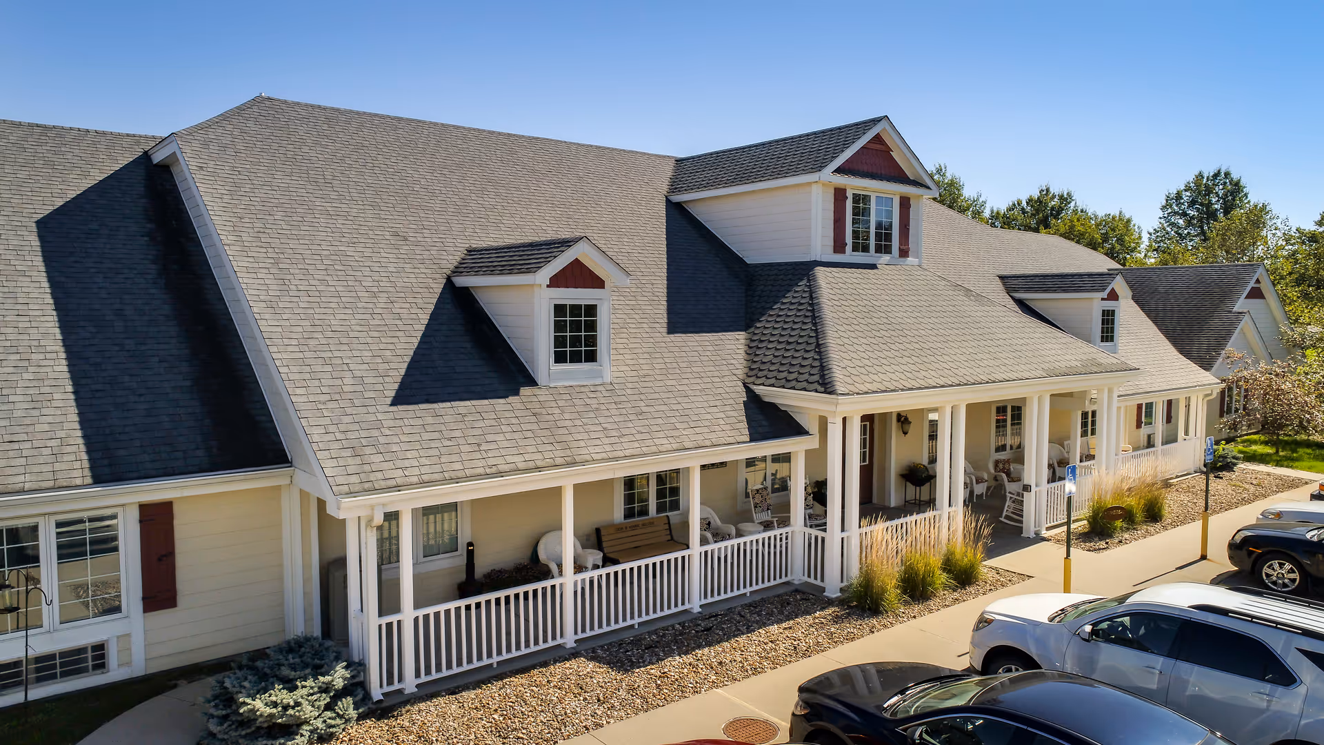 Exterior view of a single-story assisted living facility building with a gray shingled roof, white siding, and a covered porch with white railings. Several chairs and benches are placed on the porch. There are parked cars in front of the building and trees in the background under a clear sky.