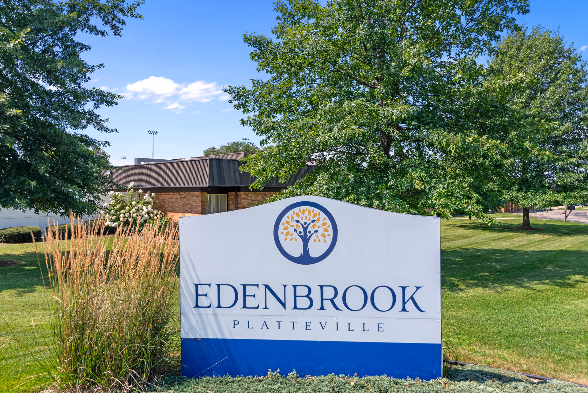 Outdoor view of the Edenbrook Platteville facility sign surrounded by green grass, trees, and plants with a building partially visible in the background under a blue sky with some clouds.