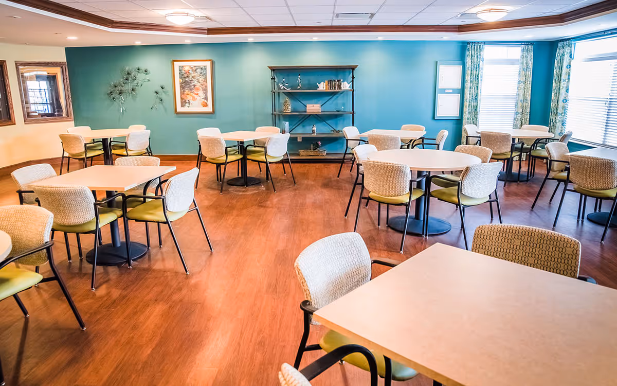 A bright dining room with multiple square and round tables surrounded by cushioned chairs. The room has wooden flooring, teal and beige walls, and large windows with patterned curtains allowing natural light to enter. There is a decorative shelf with books and ornaments against the teal wall, along with framed artwork and wall decor.