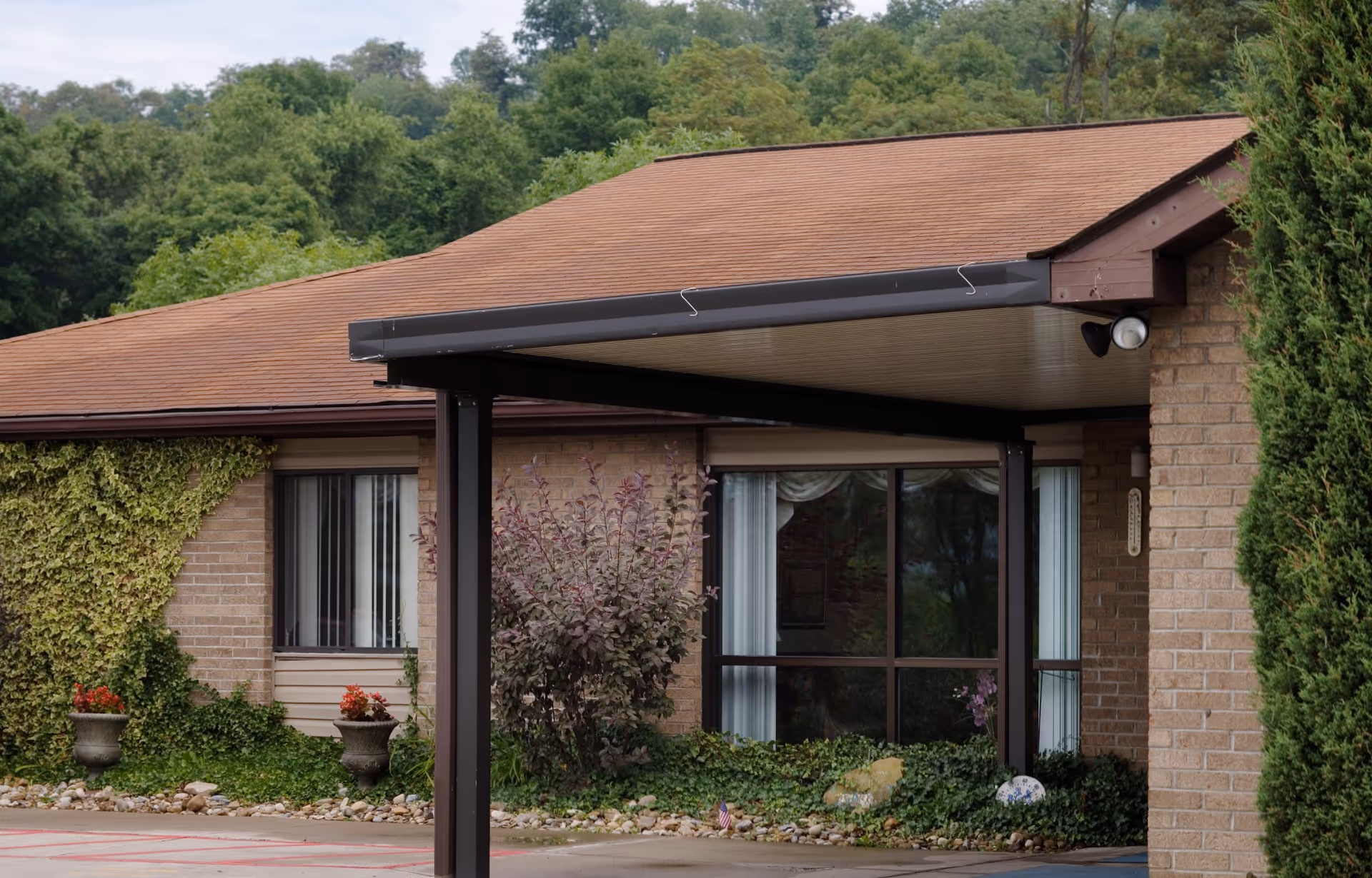 Covered entrance/portico of a brick senior living building with large windows, potted plants, shrubs, and a wooded background.