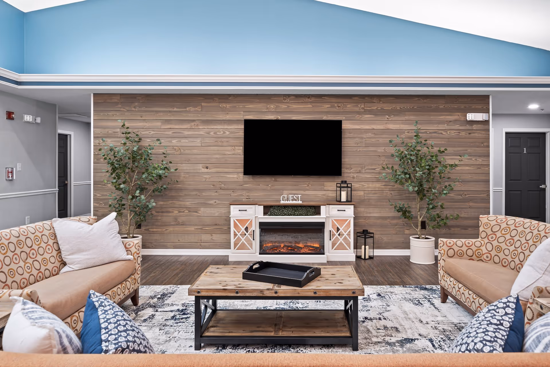 A cozy living room with two patterned sofas facing a wooden coffee table. The back wall is covered with horizontal wooden planks and features a mounted flat-screen TV above a white console with a built-in electric fireplace. Two large potted plants flank the console. The ceiling is painted light blue with white trim, and the floor has a patterned area rug over wood flooring.