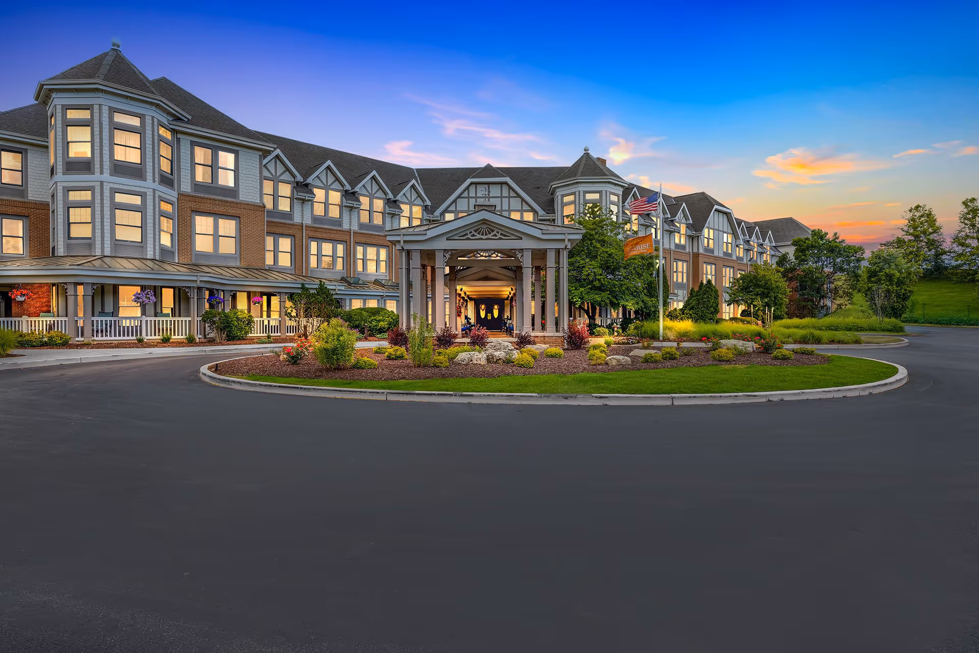Front exterior view of Sunrise of Des Peres senior living facility at sunset, showing a large multi-story building with many windows, a covered entrance with columns, landscaped garden beds, and flags near the entrance.
