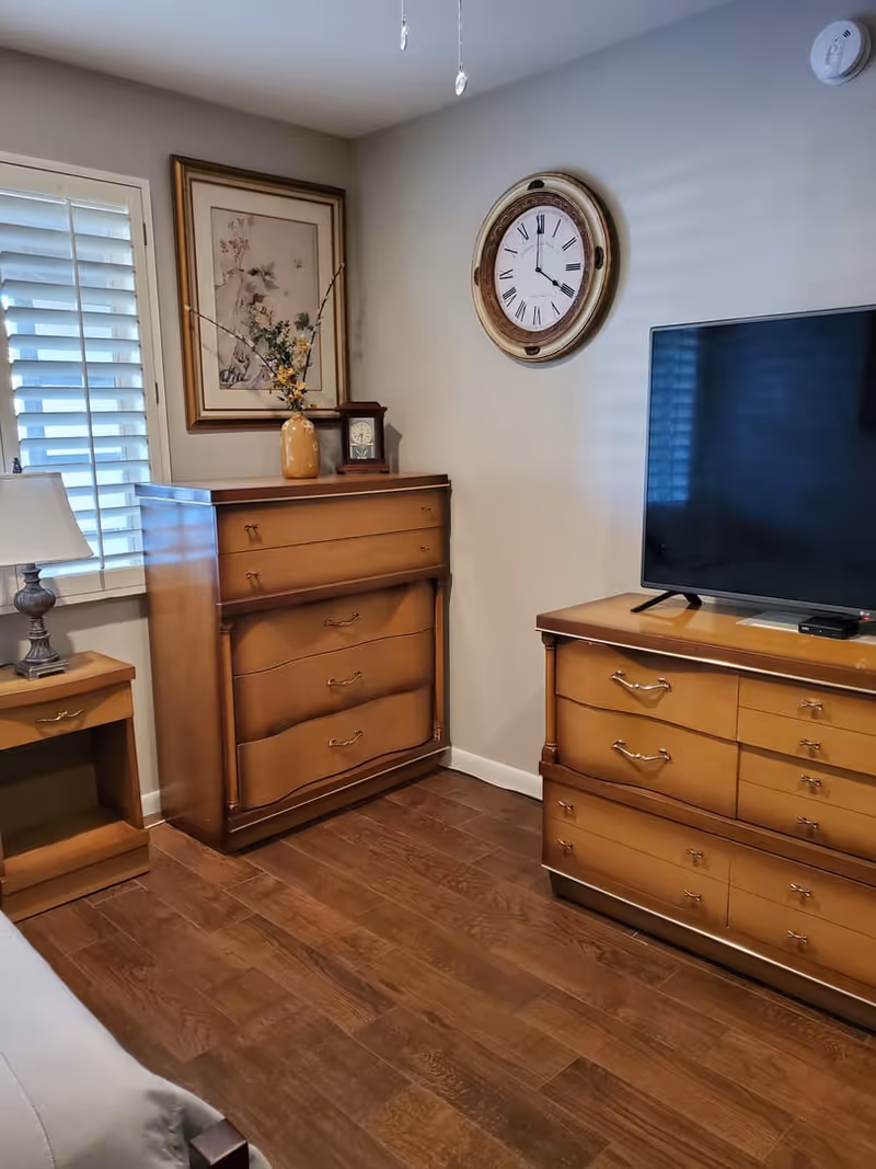 A corner of a bedroom featuring wooden furniture including a dresser with a flat-screen TV on top, a chest of drawers with a decorative vase and clock, a nightstand with a lamp, a framed floral artwork on the wall, a round wall clock, and a window with white plantation shutters. The floor is wooden and the walls are painted light gray.