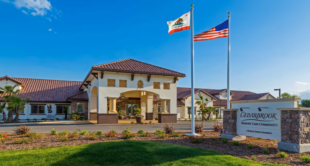 Exterior view of Cedarbrook Memory Care Community building with a tiled roof, two flagpoles displaying the California state flag and the United States flag, landscaped garden with flowers and shrubs, and a clear blue sky.
