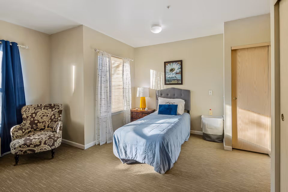 A bright and tidy bedroom with a single bed covered in a light blue comforter and a dark blue pillow. The bed has a tufted gray headboard and is positioned against a beige wall with a framed floral artwork above it. Next to the bed is a wooden nightstand with a yellow lamp and a window with sheer white curtains letting in natural light. To the left, there is a patterned armchair near a window with dark blue curtains. A beige carpet covers the floor, and a wooden sliding door is visible on the right side of the room.