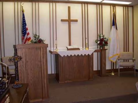 A small chapel altar with a cross, podiums, flags, and floral arrangements.