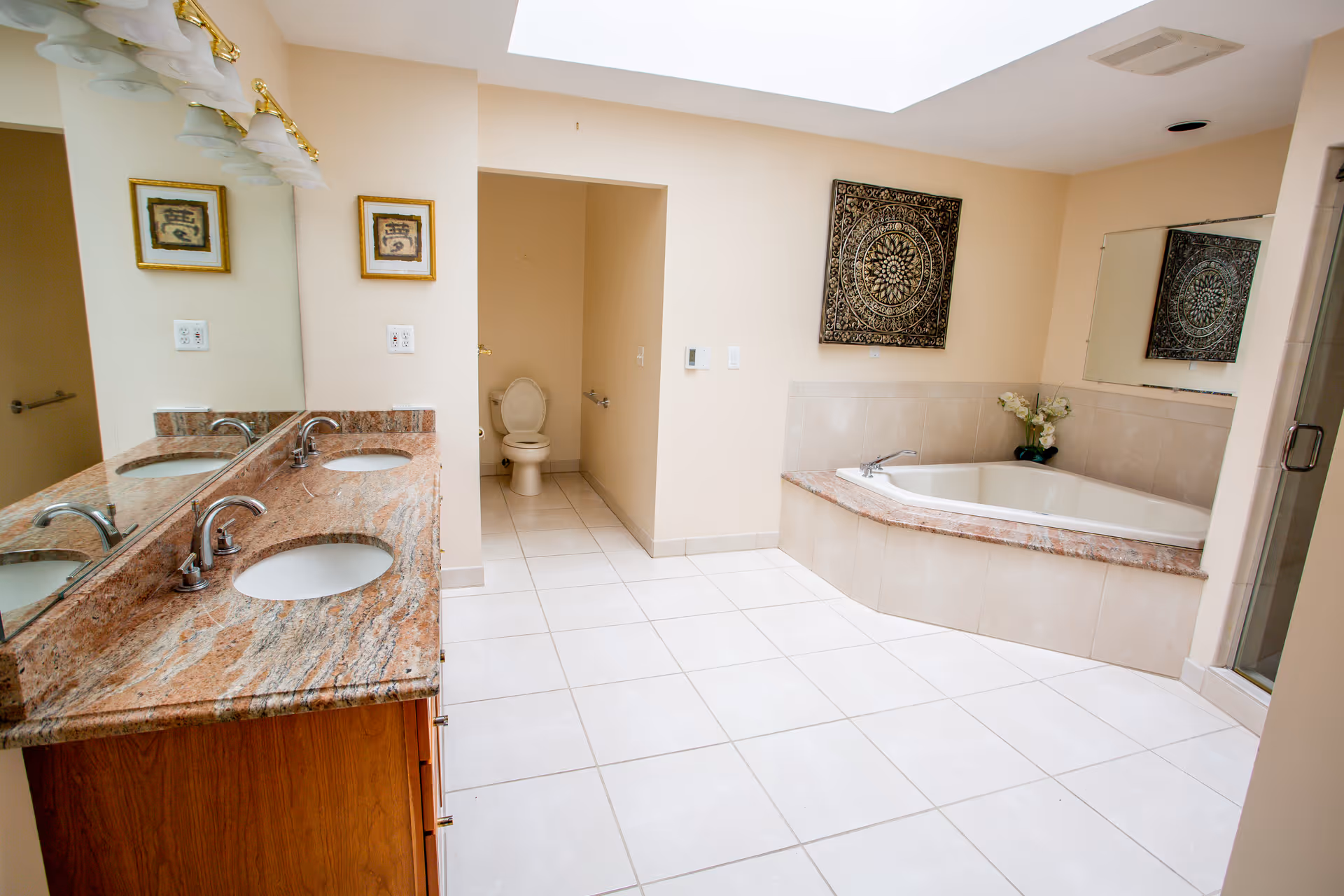 A spacious bathroom with a double sink vanity featuring a granite countertop and wooden cabinets. There is a large mirror above the sinks and two framed artworks on the wall. The bathroom includes a corner bathtub with a tiled surround, a separate enclosed shower, and a toilet in a small alcove. The floor is tiled in white, and the walls are painted a light beige color. A decorative wall hanging and a vase with flowers are placed near the bathtub.