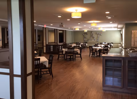 Interior view of a dining room with wooden floors, multiple tables covered with white tablecloths, and dark wooden chairs. The back wall has large decorative letters spelling 'EAT'. Ceiling lights illuminate the space warmly.
