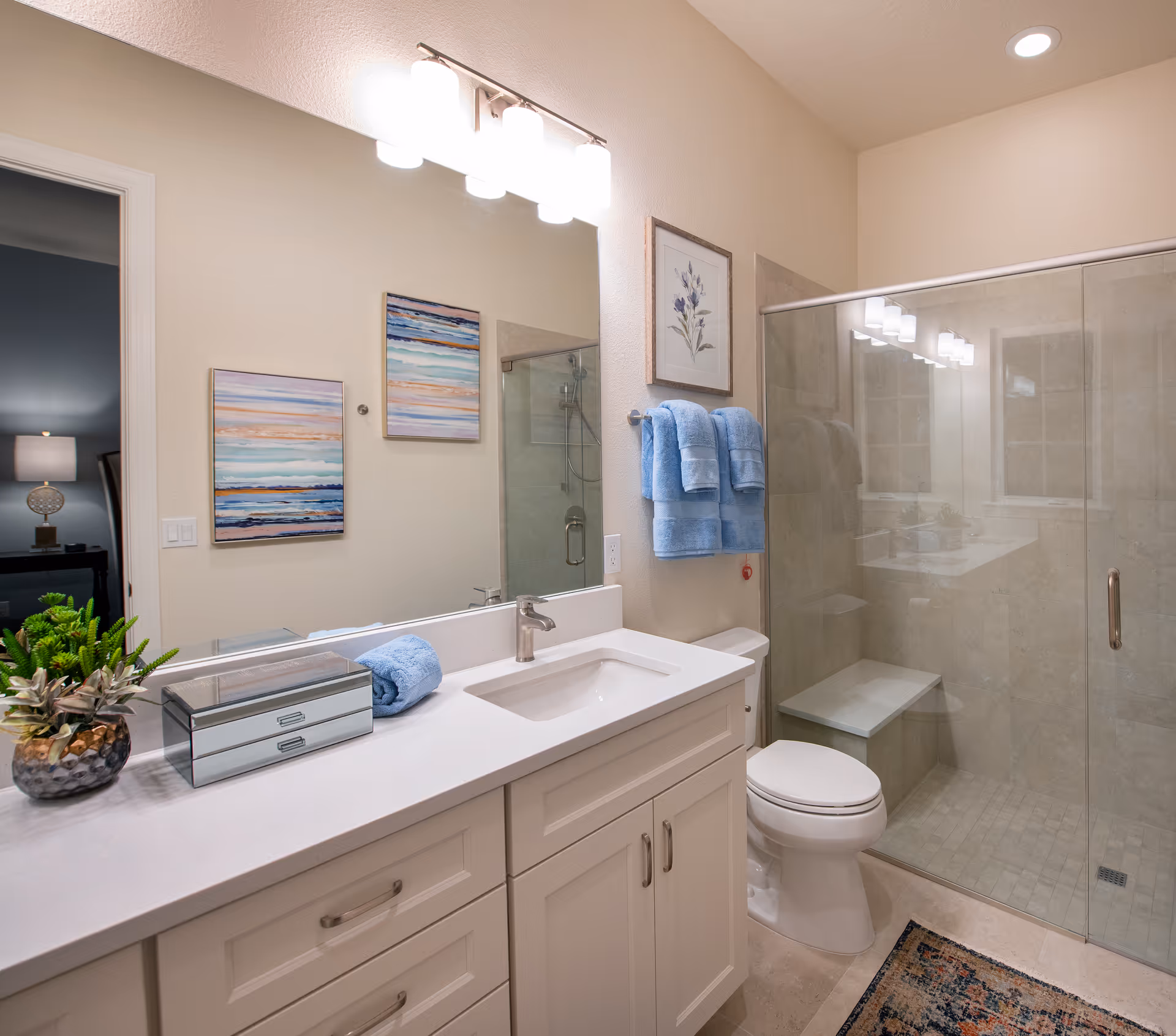A clean and modern bathroom featuring a white vanity with a sink, a large mirror with three light fixtures above it, two blue towels hanging on a towel rack, a glass-enclosed shower with a built-in bench, a toilet, and decorative artwork on the wall. There is also a small plant and a mirrored box on the vanity countertop.