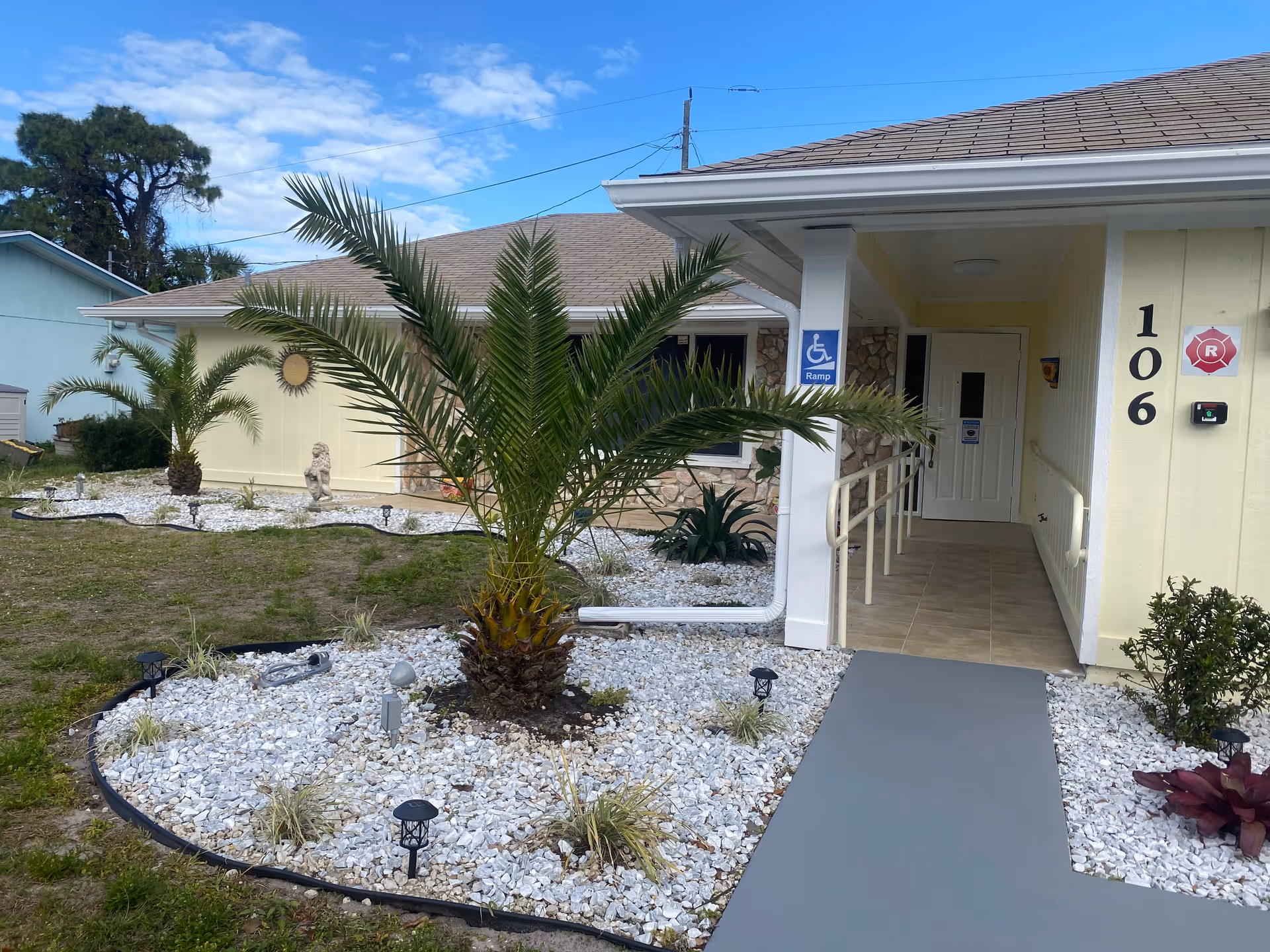 Exterior view of a single-story building with a beige and stone facade, featuring a wheelchair accessible ramp leading to the entrance. The building number 106 is displayed on the wall near the door. The landscaped front yard has palm trees, white gravel, and small plants under a partly cloudy blue sky.