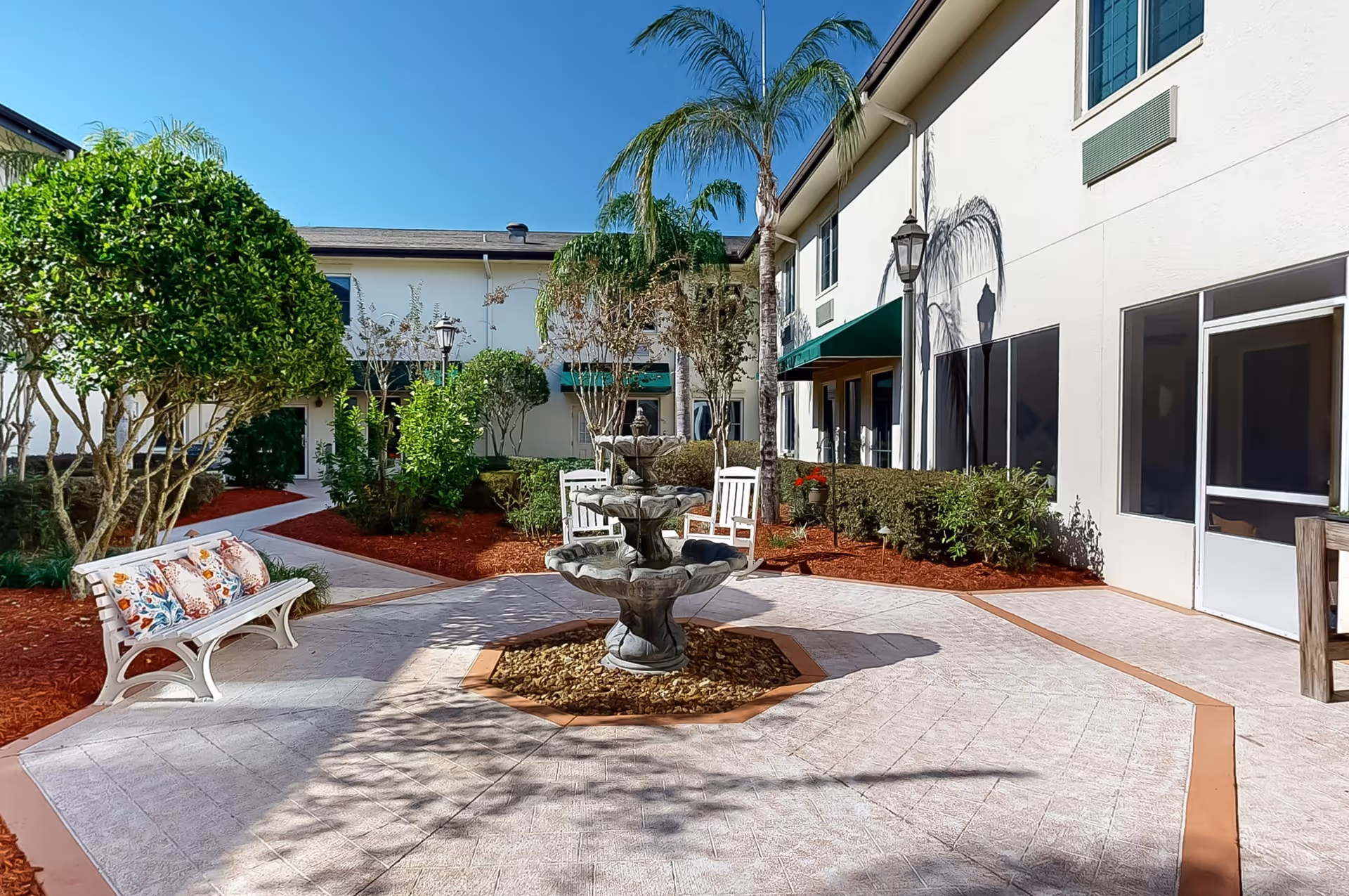 Outdoor courtyard area at The Windsor Of Ocala featuring a multi-tiered stone fountain in the center, surrounded by paved walkways, trimmed bushes, palm trees, a white bench with colorful cushions, and a two-story building with windows and green awnings under a clear blue sky.