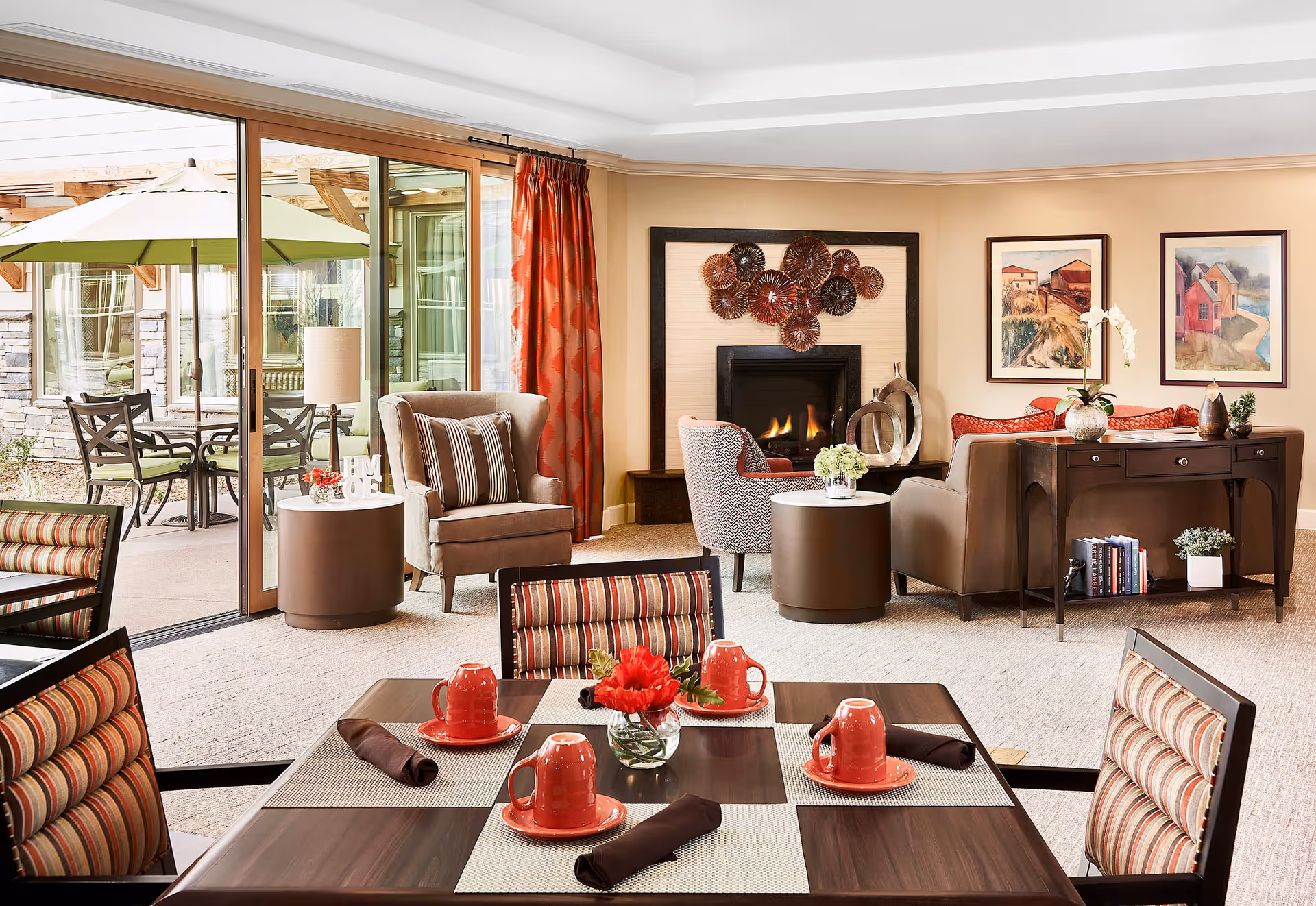 A cozy senior living common area with a dining table set for four with red cups and plates, and brown napkins. The room features comfortable armchairs, a fireplace with decorative art above it, and a console table with books and plants. Large sliding glass doors open to an outdoor patio with a table and umbrella.