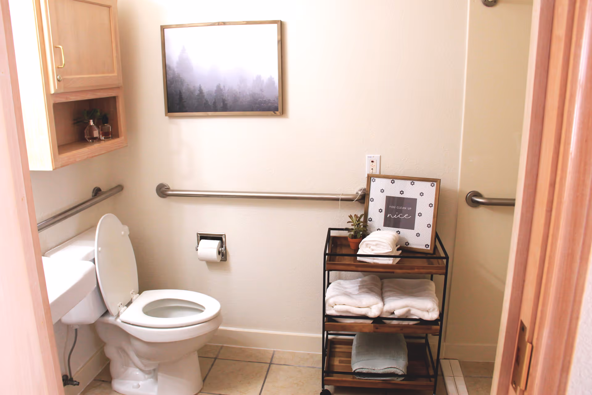 A clean bathroom with a white toilet, grab bars on the walls, a wooden cabinet, a small cart with folded towels and a framed sign, and a framed picture of a foggy forest on the wall.
