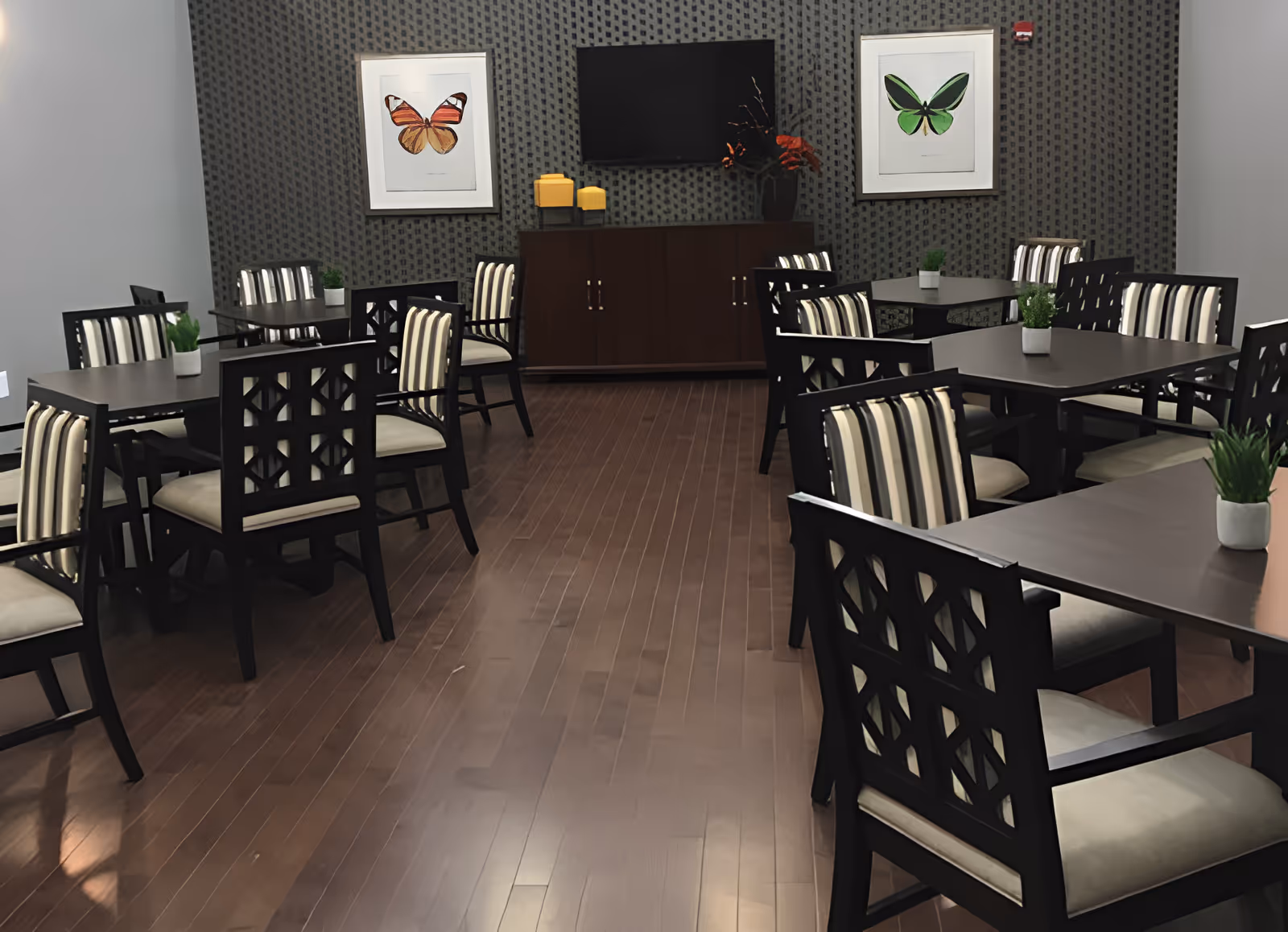 A dining room with several dark wood tables and chairs with striped cushions. The floor is wooden, and the walls have a patterned wallpaper. Two framed butterfly artworks hang on the wall, with a flat-screen TV mounted between them above a dark wood cabinet decorated with yellow candles and a vase with orange flowers. Small potted plants are placed on the tables.