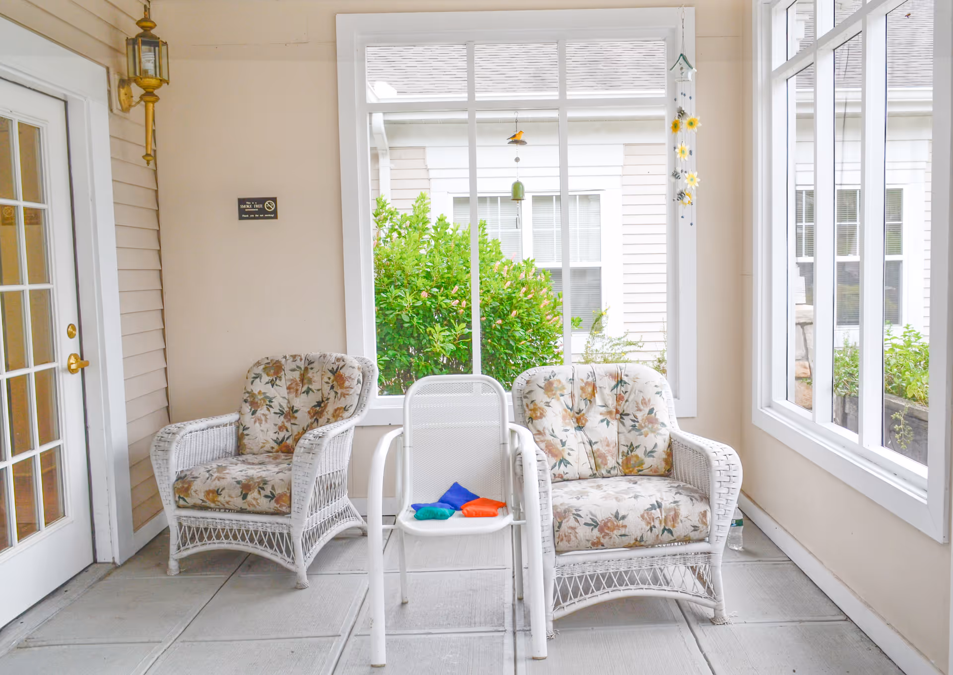 A bright enclosed porch area with two white wicker armchairs featuring floral cushions and a white metal chair in the center with colorful bean bags on the seat. Large windows show green bushes outside, and there is a hanging decoration with sunflowers near the window.