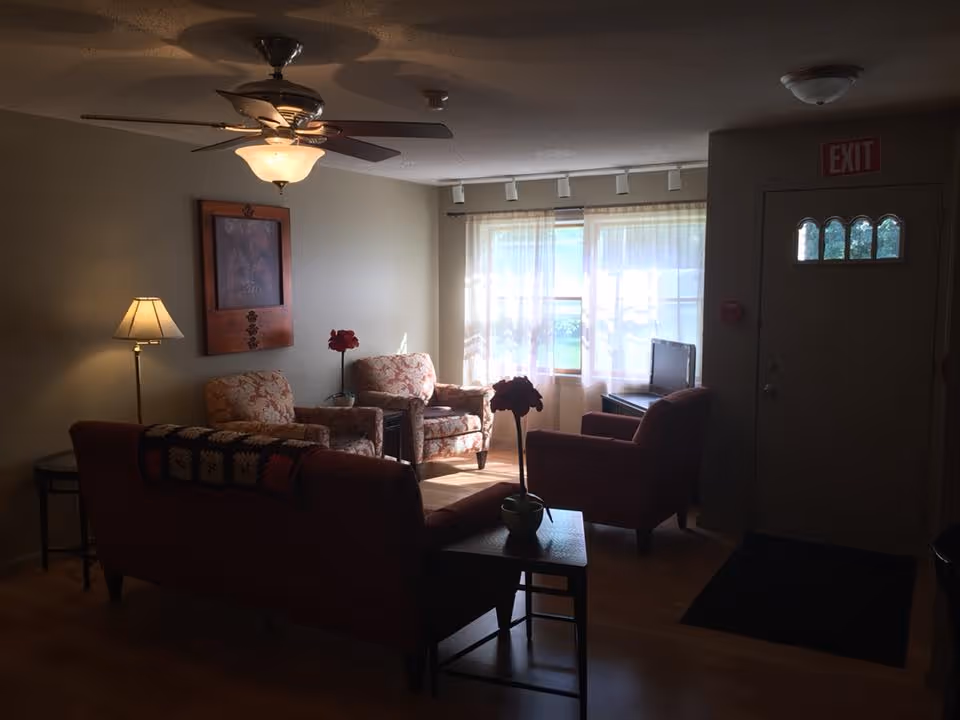 A cozy living room with a ceiling fan and light fixture, a floor lamp, two floral armchairs, a sofa with a crocheted blanket draped over the back, a small table with a potted plant, and a window with sheer curtains letting in natural light. There is an exit door with a small window and an exit sign above it.