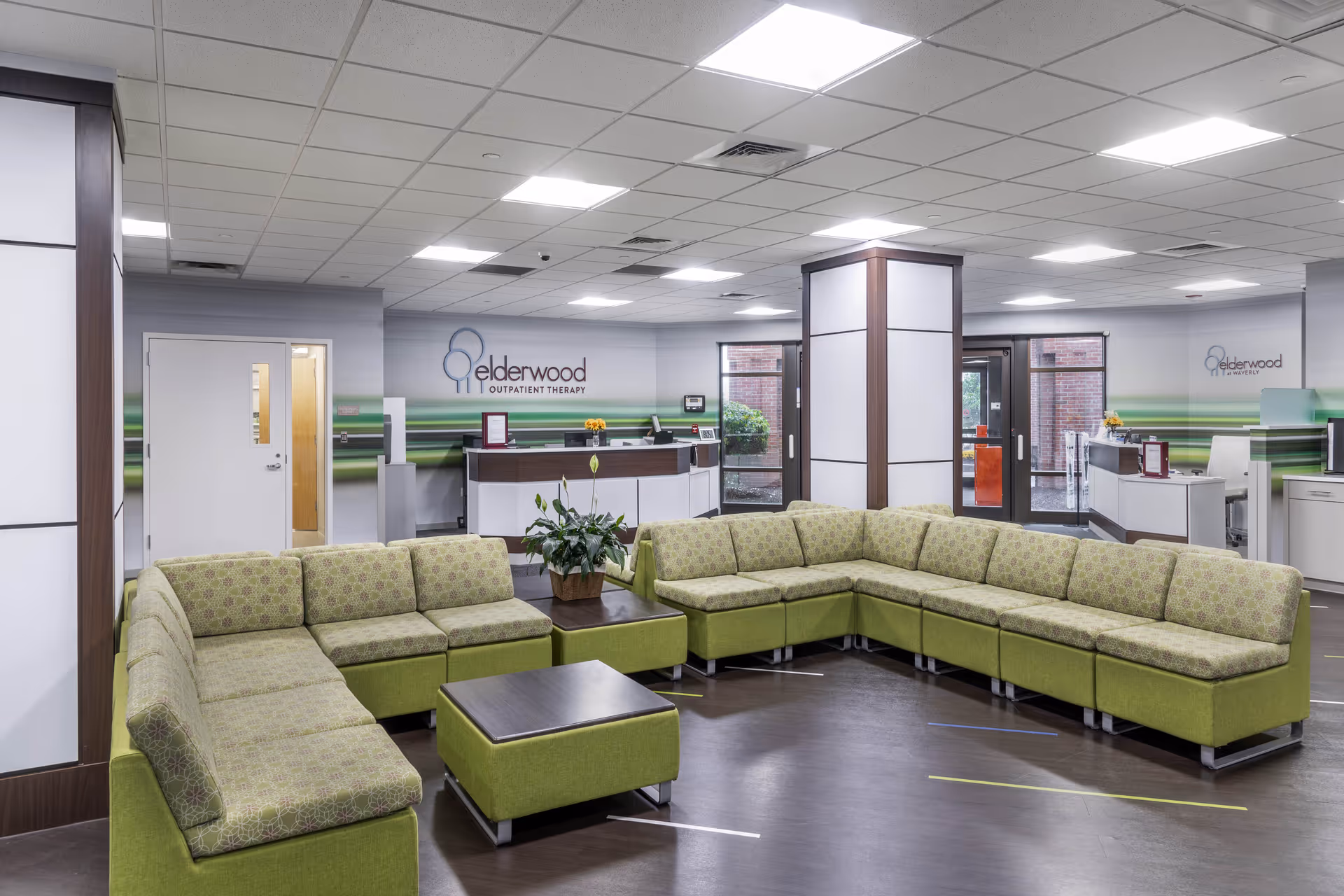 A spacious waiting area with a large green sectional sofa arranged around a central coffee table and ottoman. The room has a modern design with a drop ceiling and bright lighting. In the background, there is a reception desk with the Elderwood Outpatient Therapy logo on the wall. Glass doors and windows provide natural light and a view of the outside.