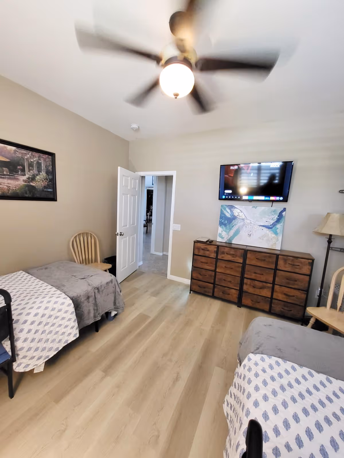 A bedroom with two single beds covered with gray and white patterned blankets, a wooden chair, a wooden dresser with multiple drawers, a flat-screen TV mounted on the wall above the dresser, a floor lamp, a ceiling fan with a light, and a painting on the wall. The room has light-colored wooden flooring and beige walls.