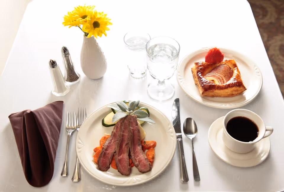 A neatly set dining table with a plate of sliced steak on vegetables, a plate with a pastry topped with a strawberry slice, a cup of black coffee on a saucer, two glasses of water, a folded brown napkin, silverware, salt and pepper shakers, and a small white vase with yellow flowers.