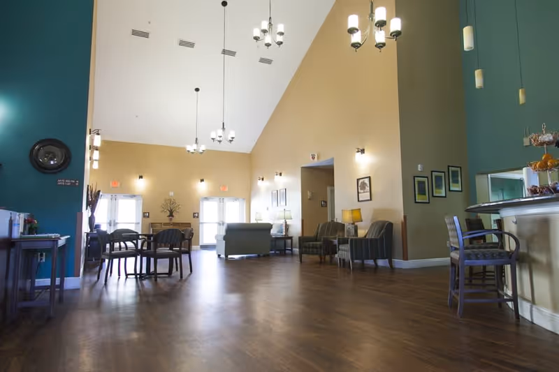 Spacious senior living facility common area with high vaulted ceilings, multiple chandeliers, wooden flooring, and seating arrangements including chairs and sofas. The walls are painted in beige and teal colors, with framed pictures and lamps placed around the room. There is a counter with chairs on the right side and double doors at the far end letting in natural light.