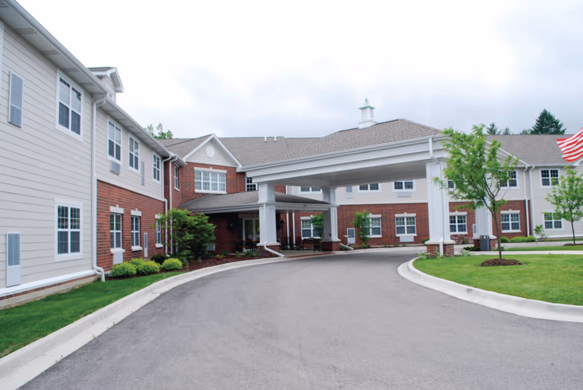 Entrance driveway and covered porte-cochère of a two-story brick-and-siding senior living building with landscaping.