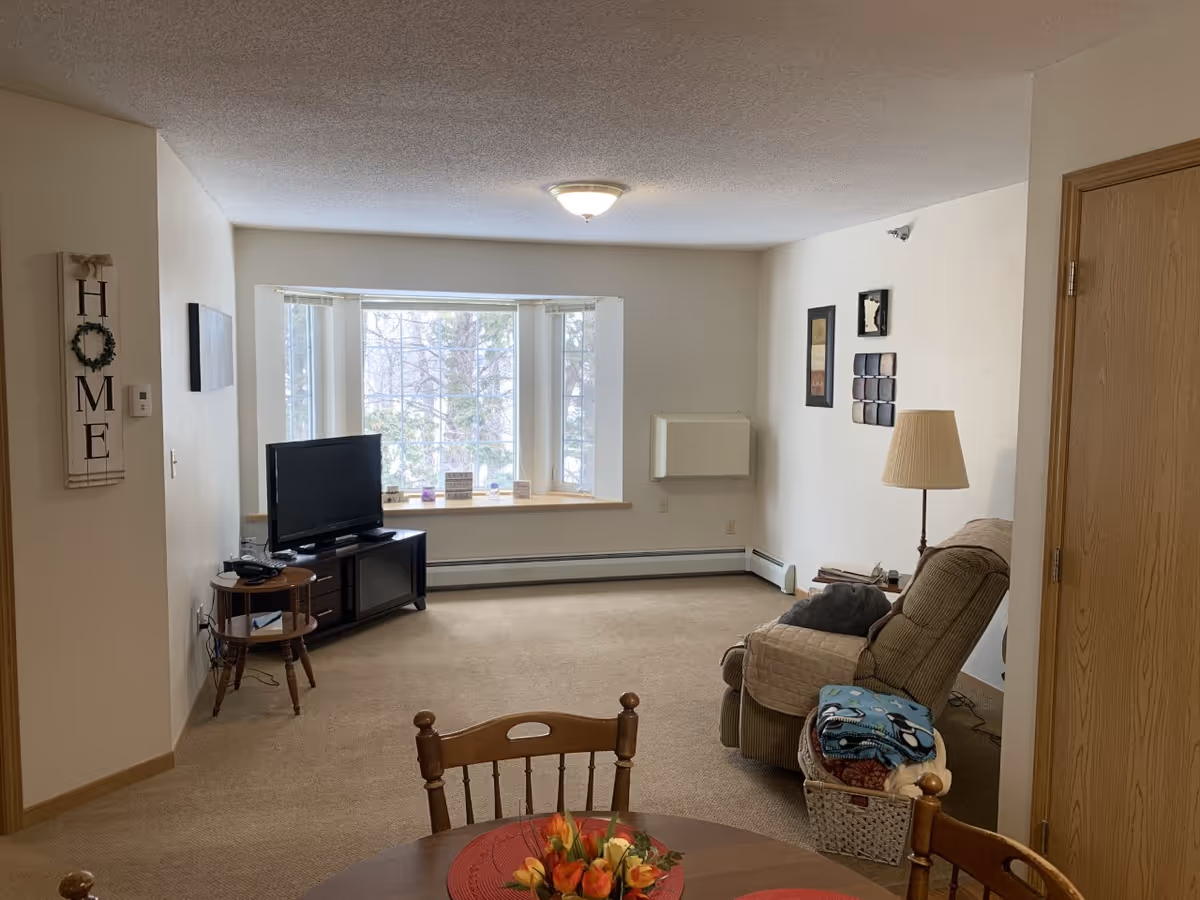 A cozy living room with beige carpet and white walls. There is a large window with a wooden sill letting in natural light. A flat-screen TV is placed on a dark wooden TV stand in the corner. A comfortable recliner chair with a blanket and a basket of folded blankets next to it is positioned near a floor lamp. A round wooden dining table with chairs and a floral centerpiece is partially visible in the foreground. Wall decorations include a vertical 'HOME' sign and framed artwork.