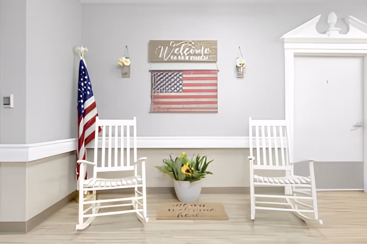A hallway corner in a senior living facility with two white rocking chairs on either side of a potted plant with yellow flowers. An American flag stands behind the left chair. On the wall above, there is a wooden sign that says 'Welcome' and a rustic American flag decoration. The floor has a welcome mat in front of a white door with decorative molding.