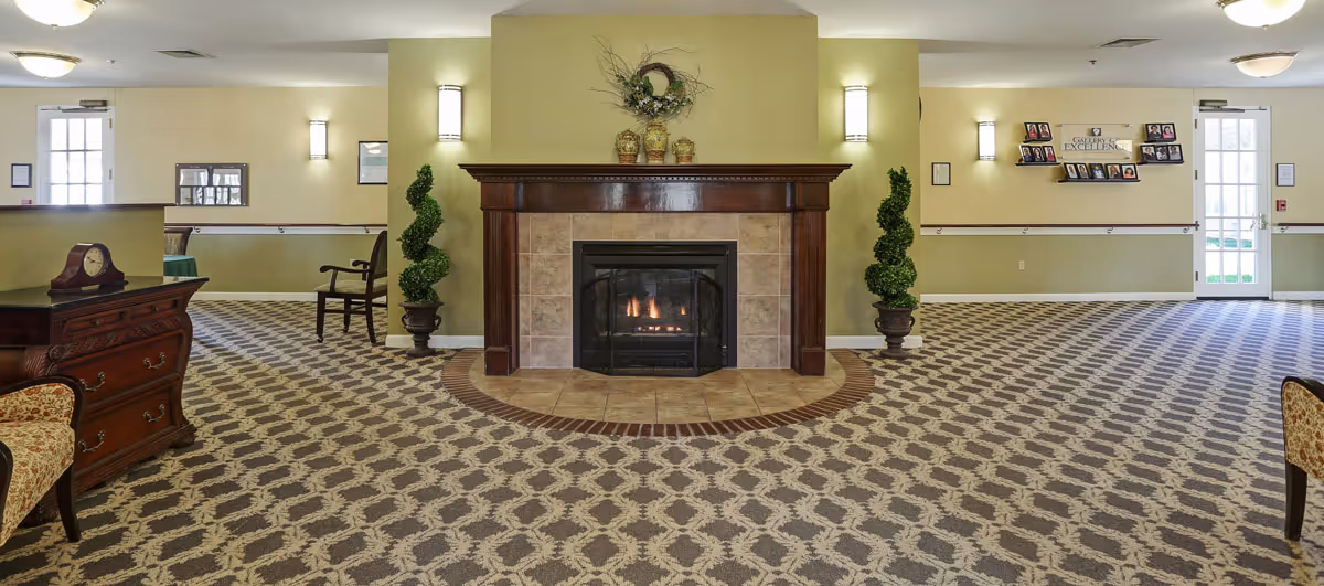 A spacious interior room with a patterned carpet, a central fireplace with a wooden mantle, two potted spiral topiary plants on either side of the fireplace, wall sconces providing lighting, framed pictures on the walls, and wooden furniture including chairs and a chest with a clock on it.