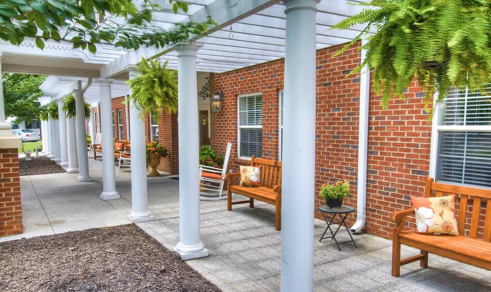 Outdoor covered walkway with white columns and hanging ferns at TerraBella Pheasant Ridge. Wooden benches with decorative pillows and small tables with potted plants line the walkway next to a brick building with windows.