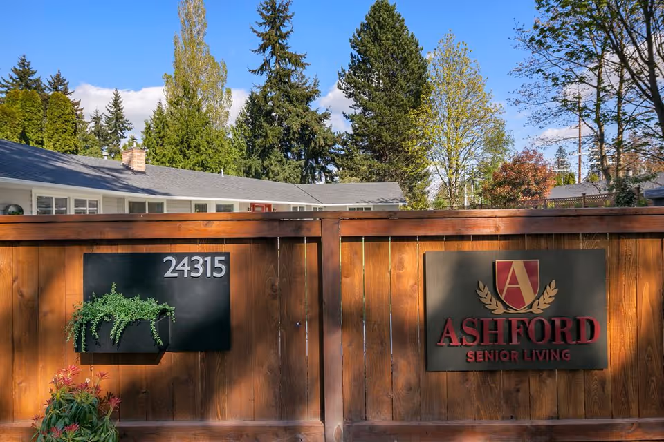 Wooden fence with two black signs mounted on it. The left sign displays the number 24315 with a small green plant attached below it. The right sign features the Ashford Senior Living logo with a stylized letter A and laurel leaves above the text. Behind the fence, a single-story building with a gray roof and white walls is partially visible, surrounded by tall trees under a blue sky with some clouds.
