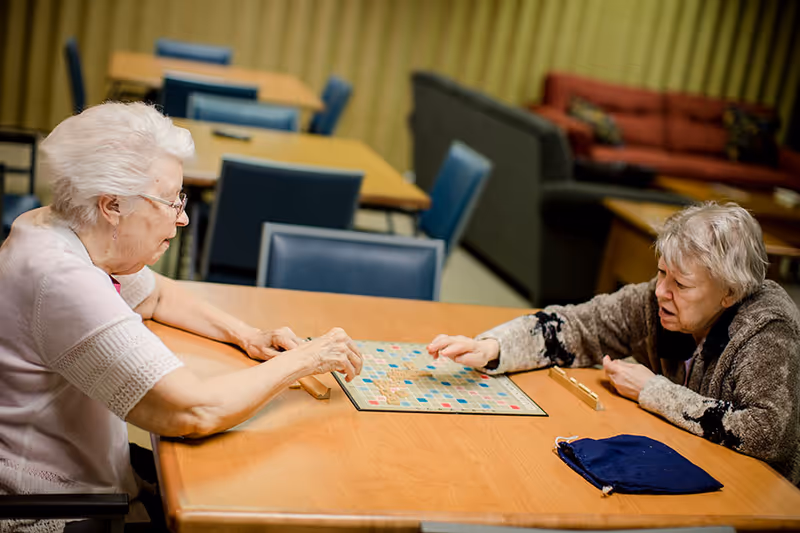 Two elderly women sitting at a wooden table playing a game of Scrabble in a room with blue chairs and a couch in the background.