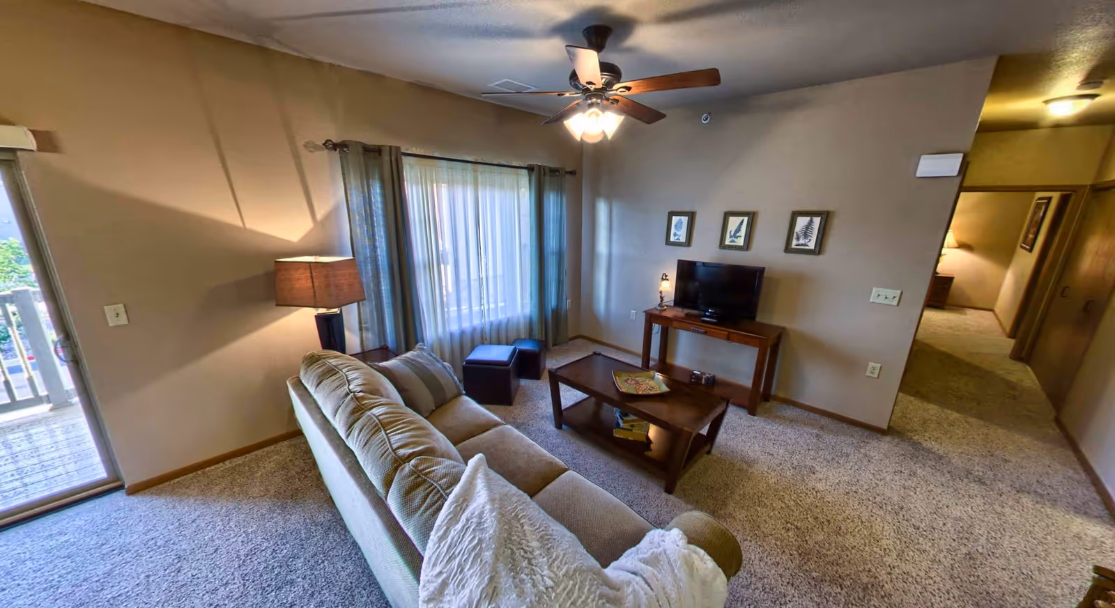 A cozy living room with a beige couch, a wooden coffee table with books and a decorative plate, a wooden TV stand with a flat-screen TV, three framed pictures on the wall, a floor lamp, and a ceiling fan with lights. There is a window with sheer curtains and a sliding glass door leading to a balcony. The room has beige walls and carpeted floors.