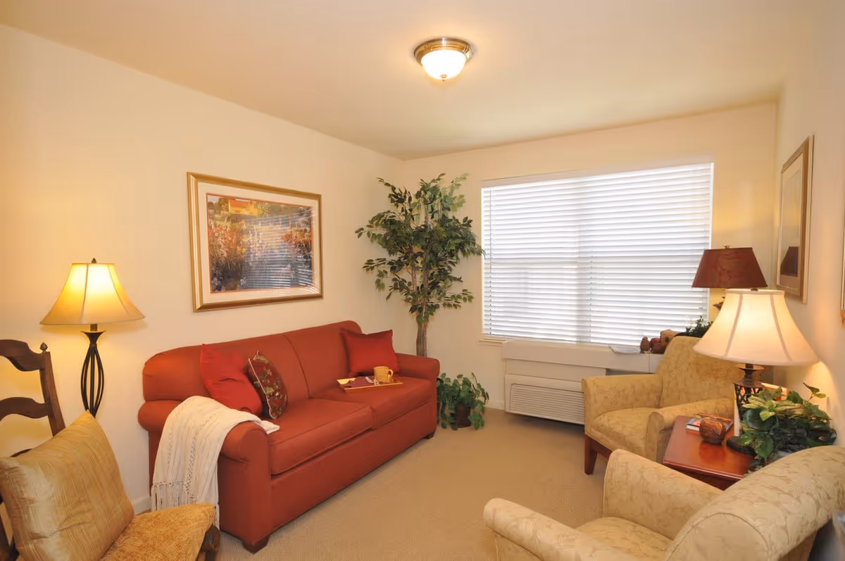 A cozy living room with a red sofa adorned with cushions and a white throw blanket, two beige armchairs, a wooden side table with a lamp and decorative plants, a large window with blinds, a framed painting on the wall, and a floor lamp beside a wooden chair.