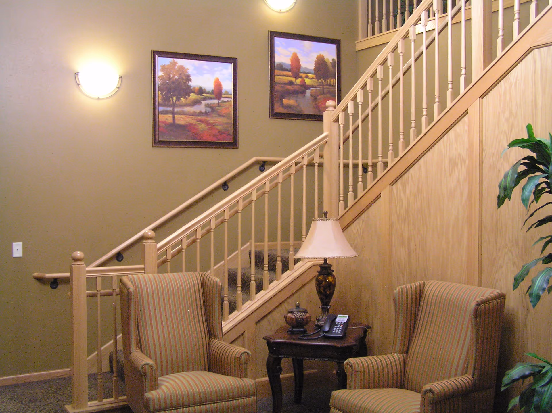 A cozy seating area with two striped armchairs and a small wooden table between them holding a decorative lamp and a telephone. Behind the seating area is a staircase with wooden railings and two framed landscape paintings on the wall. A green plant is visible on the right side.