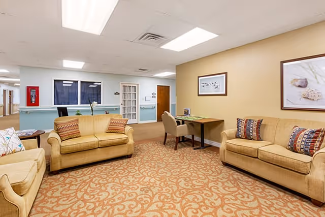 A cozy seating area in a senior living facility with three beige sofas adorned with patterned cushions arranged around a carpet with an intricate design. There is a small table with two chairs against a yellow wall, which has two framed pictures hanging above it. The background shows a hallway with a blue wall and a fire extinguisher mounted on it.