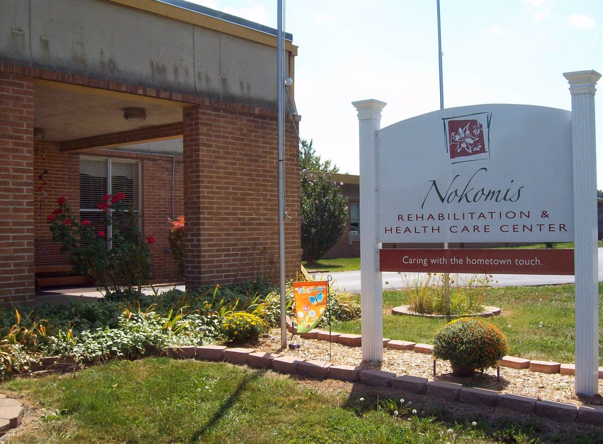 Front entrance and sign for Nokomis Rehabilitation & Health Care Center beside a landscaped lawn in front of a brick building.