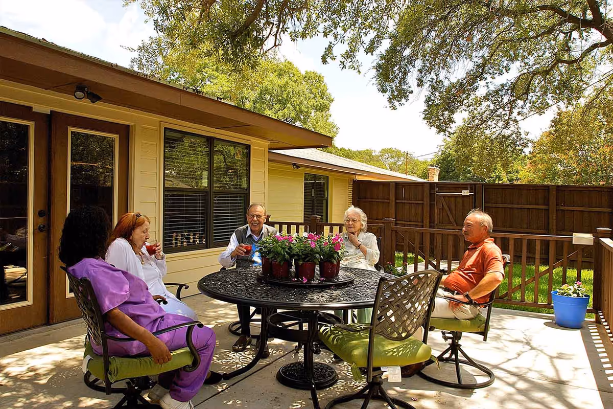 Four elderly people and a caregiver sitting around a round outdoor table with potted flowers on a patio, enjoying drinks and conversation. The patio is adjacent to a yellow building with large windows and a wooden fence surrounds the area. Trees and greenery are visible in the background.