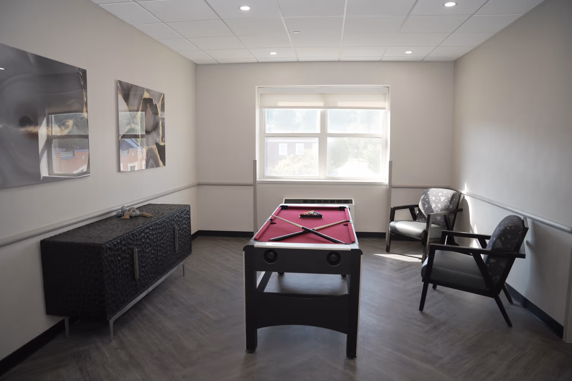 A small recreation room with a red felt pool table in the center, two chairs with patterned cushions on the right side, a black textured cabinet on the left, and two abstract art pieces hanging on the left wall. A large window at the back lets in natural light.